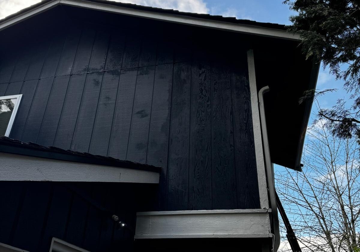 Exterior view of a two-story home with dark blue siding and white trim, showing the upper wall before siding repair under a partly cloudy sky.